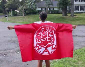 May include: A bright red flag with a large white emblem featuring Arabic script and the letters "W.A.C." is held open. The flag is displayed outdoors, with a person holding it up against a backdrop of green grass and trees.