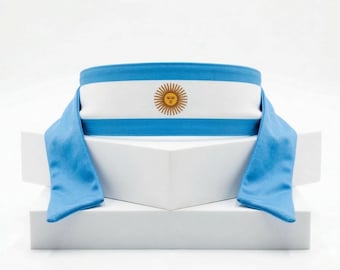 May include: A headband featuring the Argentinian flag design. The headband has a white center stripe with a gold sun emblem, bordered by light blue stripes and flowing blue fabric.