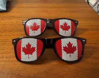 May include: Two pairs of black-framed sunglasses with the Canadian flag design on the lenses. The flag features a red maple leaf on a white background, with red borders. The sunglasses are displayed on a wooden surface.