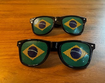 May include: Two pairs of black-framed sunglasses with the Brazilian flag design on the lenses. The flag features a green background with a yellow diamond, a blue circle, and stars.
