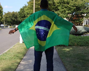 May include: A person stands outdoors, wearing a Brazilian flag cape. The cape is green with a yellow diamond, a blue circle with stars, and the words "Ordem e Progresso". The person is facing away from the camera.