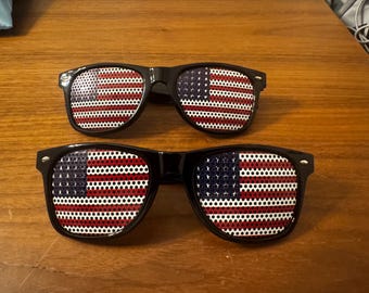 May include: Two pairs of black-framed sunglasses with the American flag design on the lenses. The lenses feature a red and white striped pattern with a blue square containing white stars. The sunglasses are displayed on a wooden surface.