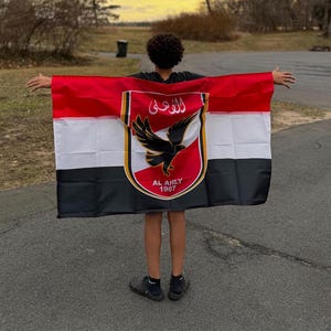 May include: A person holding a flag with red, white, and black horizontal stripes. The flag features a crest with a black eagle and Arabic text, with "AL AHLY 1907" below.