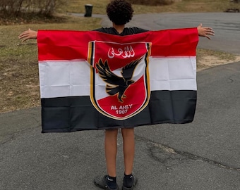 May include: A person holding a flag with red, white, and black horizontal stripes. The flag features a crest with a black eagle and Arabic text, with "AL AHLY 1907" below.