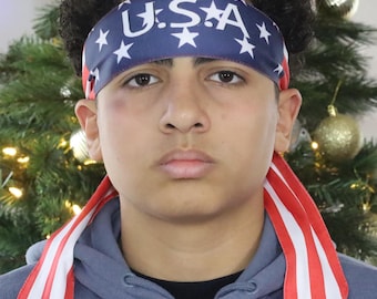 May include: A person wearing a blue headband with the word "USA" and white stars. The headband is paired with a gray hoodie and red and white striped fabric hanging from the neck. A Christmas tree is in the background.