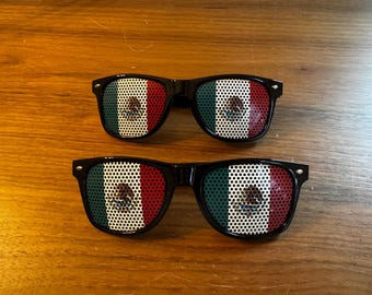 May include: Two pairs of black-framed sunglasses with the Mexican flag design on the lenses. The flag features green, white, and red vertical stripes with the Mexican coat of arms in the center. The sunglasses are displayed on a wooden surface.