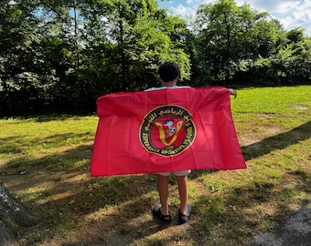 May include: A red flag with a circular emblem featuring text in Arabic and French, along with a crest. The flag is held in front of a person standing in a grassy area with trees in the background.