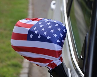 May include: A car side mirror cover featuring the American flag design. The cover displays red and white stripes with a blue field of white stars. The cover is attached to a car's side mirror, with a green background.