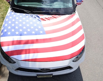 May include: A white car with a hood cover featuring the American flag design. The flag has red and white stripes and a blue field with white stars. Small American flags are attached to the side mirrors.