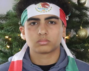 May include: A white, green, and red headband with the Mexican coat of arms in the center. The headband is tied at the back and is worn by a person. The ends of the headband are in the colors of the Mexican flag.