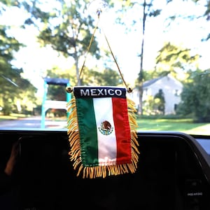 May include: A small, decorative Mexican flag with the word "MEXICO" at the top. The flag features green, white, and red vertical stripes, with the Mexican coat of arms in the center. Gold fringe adorns the bottom.