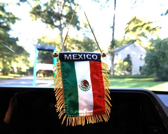 May include: A small, decorative Mexican flag with the word "MEXICO" at the top. The flag features green, white, and red vertical stripes, with the Mexican coat of arms in the center. Gold fringe adorns the bottom.