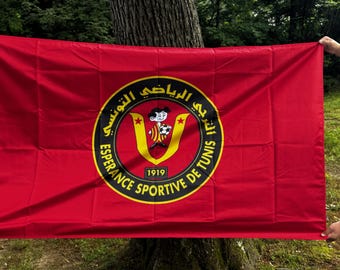 May include: A red flag featuring the emblem of Espérance Sportive de Tunis. The emblem is a yellow circle with black text and a cartoon figure holding a soccer ball. The flag also has the text "1919".