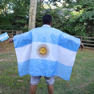 May include: A person wearing a light blue and white Argentina flag poncho with a sun emblem. The person is also holding a small Argentina flag. The background includes green grass, trees, and a wooden fence.