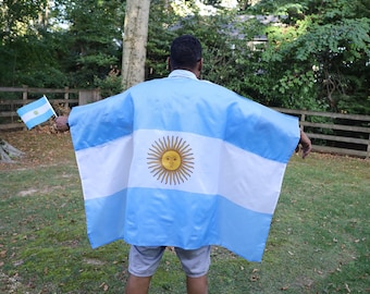 May include: A person wearing a light blue and white Argentina flag poncho with a sun emblem. The person is also holding a small Argentina flag. The background includes green grass, trees, and a wooden fence.