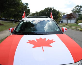 May include: A car decorated with the Canadian flag, featuring a large red maple leaf on a white background. The hood, side mirrors, and roof are covered in the flag design. Two Canadian flags are mounted on the roof.