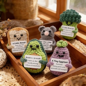 Può includere: Una collezione di alimenti all'uncinetto fatti a mano, tra cui una fetta di pane, un avocado, un topo, un broccolo e una melanzana. Ogni articolo ha un piccolo cartello con un messaggio motivazionale. Gli articoli sono esposti in un vassoio di legno.