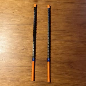 Könnte beinhalten: Zwei schwarze und orangefarbene Drumsticks mit blauen Akzenten. Die Drumsticks sind lang und dünn, mit einem strukturierten schwarzen Griff und orangefarbenen Spitzen. Sie liegen auf einer Holzoberfläche.