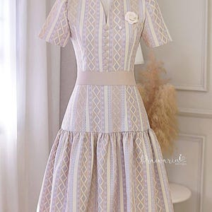 May include: A light purple and beige patterned dress with a white flower brooch. The dress has a V-neckline, short sleeves, and a fitted bodice. The skirt is tiered and falls to just above the knee.