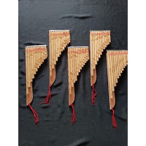 Puede incluir: Cinco flautas de pan de bambú, dispuestas sobre una superficie negra. Cada flauta tiene una serie de tubos graduados, una base de madera y una cuerda roja. Las flautas son de color bambú natural con detalles rojos.