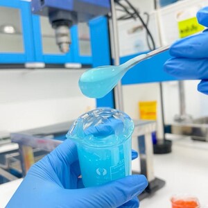 May include: A laboratory setting with a beaker containing a light blue liquid. A stainless steel spoon holds a portion of the same liquid above the beaker. The scene is viewed from a low angle, with blue gloves visible.