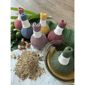 Puede incluir: Una colección de bolas de compresión herbales redondas y coloridas en tonos rosa, azul, amarillo y burdeos, con mangos envueltos en blanco. Las bolas están dispuestas con hierbas, especias y una bola de compresión verde remojándose en un cuenco de vidrio.