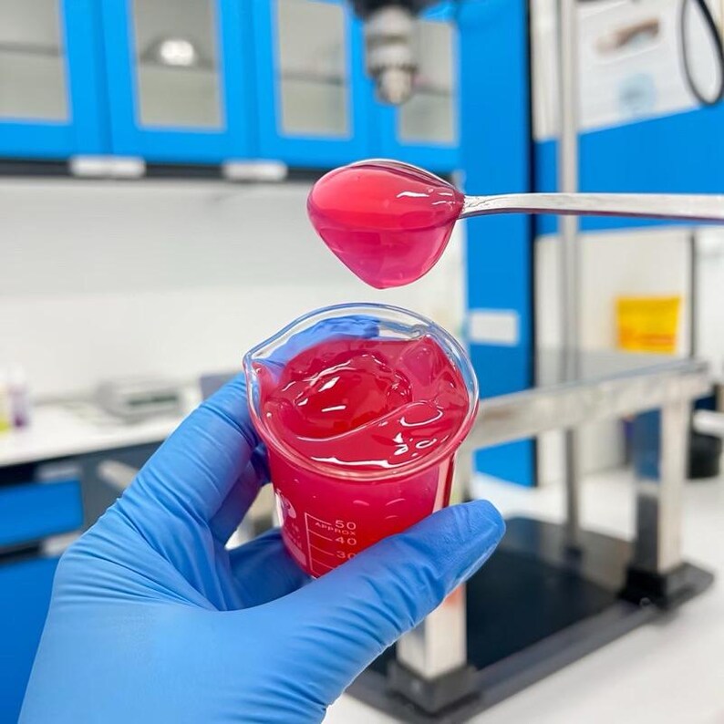 May include: A close-up shot of a beaker filled with a vibrant pink gel. A spoonful of the same gel is held above the beaker. The person holding the beaker is wearing a blue glove. The background features blue cabinets and laboratory equipment.