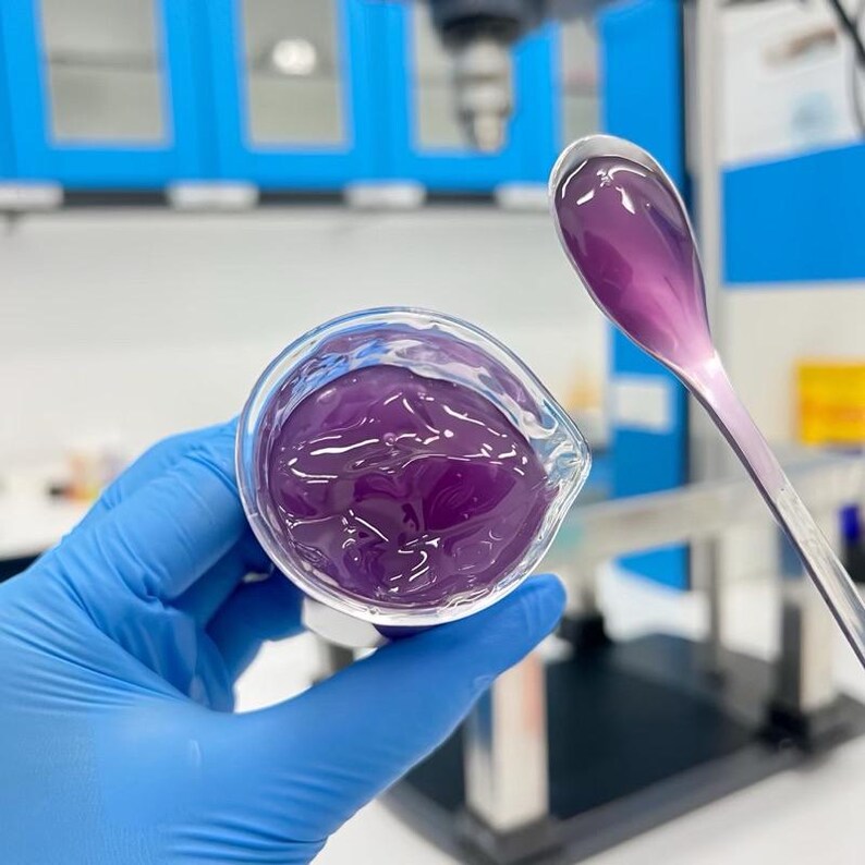 May include: A close-up of a clear glass beaker filled with a vibrant purple gel. A matching purple gel is on a spoon held nearby. The beaker is held by a gloved hand, with a laboratory setting in the background.