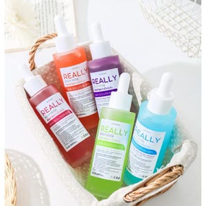 May include: A woven basket holds five glass bottles of skincare products. The bottles are filled with colorful liquids in shades of red, orange, purple, green, and blue. Each bottle has a white dropper and a label that reads "REALLY BODY DOSE AHA SOLUTION 90%."