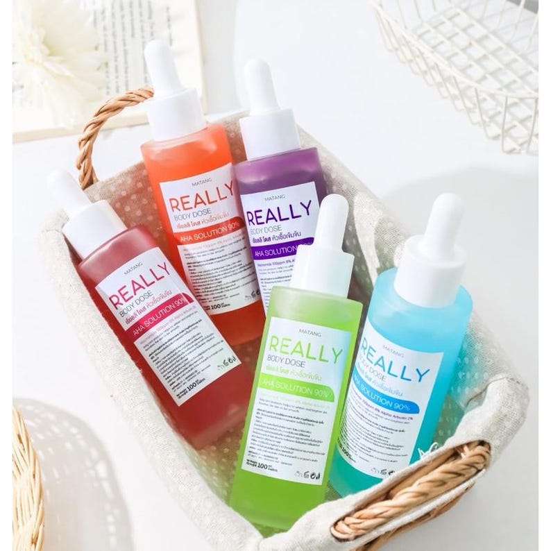 May include: A woven basket holds five glass bottles of skincare products. The bottles are filled with colorful liquids in shades of red, orange, purple, green, and blue. Each bottle has a white dropper and a label that reads "REALLY BODY DOSE AHA SOLUTION 90%."