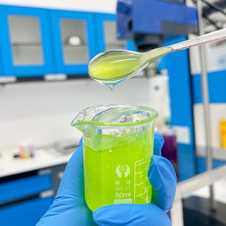 May include: A beaker filled with 50ml of bright green liquid is held by a gloved hand. A spoon-like tool holds a drop of the same liquid above the beaker. The laboratory setting includes blue cabinets and equipment.