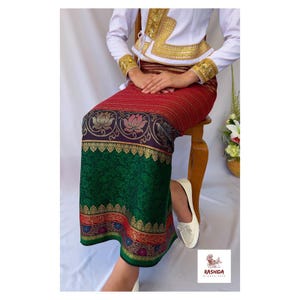 Puede incluir: Una falda tradicional con un patrón rojo y verde. La falda tiene un diseño de flor de loto y adornos dorados. La parte superior es blanca con detalles dorados. El atuendo se usa con zapatos blancos. La marca "RASHIDA" es visible.