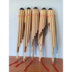 May include: Five traditional reed panpipes made of bamboo. Each instrument has a black top and is tied with a red string. The pipes are arranged vertically on a wooden surface.