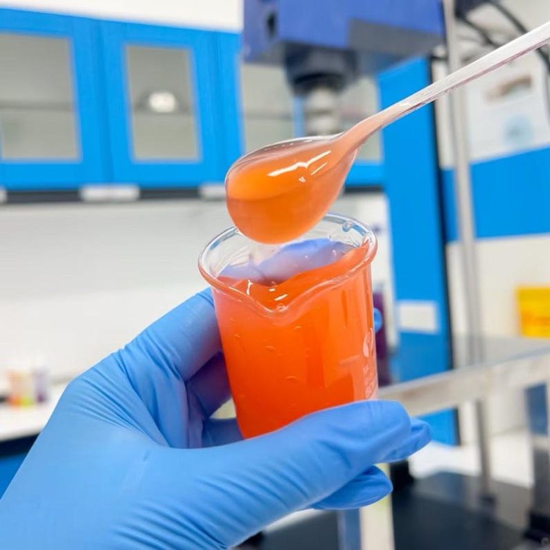 May include: A clear beaker filled with a bright orange liquid, with a spoonful of the same liquid being poured into it. The liquid is viscous and appears to be a chemical solution. The background shows a laboratory setting with blue cabinets and equipment.
