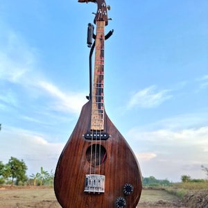 Puede incluir: Un Phin de madera hecho a mano, un instrumento de cuerda tailandés tradicional. El instrumento presenta un clavijero tallado en madera, un cuerpo en forma de lágrima y un puente con perillas de afinación. La veta de la madera es visible.