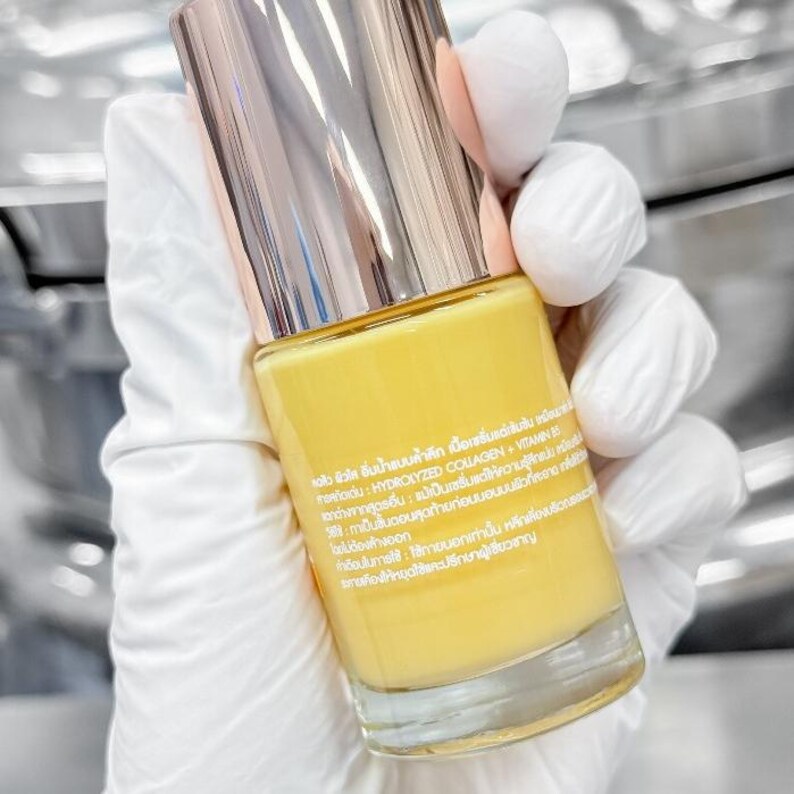 May include: A yellow cosmetic serum bottle with a metallic gold cap and a clear glass base. The bottle is held by a person wearing white gloves. The label contains text in a foreign language.