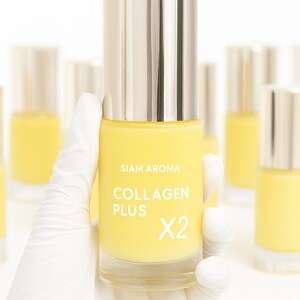 May include: A yellow bottle of "Collagen Plus X2" serum is held by a gloved hand. The bottle has a silver cap and the brand name "SIAM AROMA" is printed on the front. Other bottles are visible in the background.