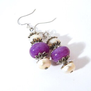 May include: A pair of dangle earrings featuring purple and cream-colored beads. The earrings have silver-colored hooks and decorative metal accents. The beads are oval-shaped and arranged in a vertical pattern, creating a stylish accessory.