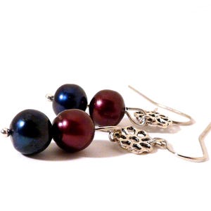 May include: A pair of dangle earrings featuring dark blue and burgundy-colored pearls. The earrings have silver-colored, openwork floral accents and curved ear wires. The jewelry is set against a white background.