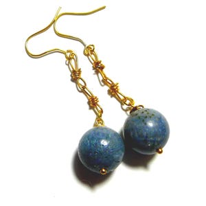 May include: A pair of dangle earrings featuring blue spherical beads. The beads are suspended from gold-colored wire chains and gold-colored ear wires. The earrings are set against a white background.