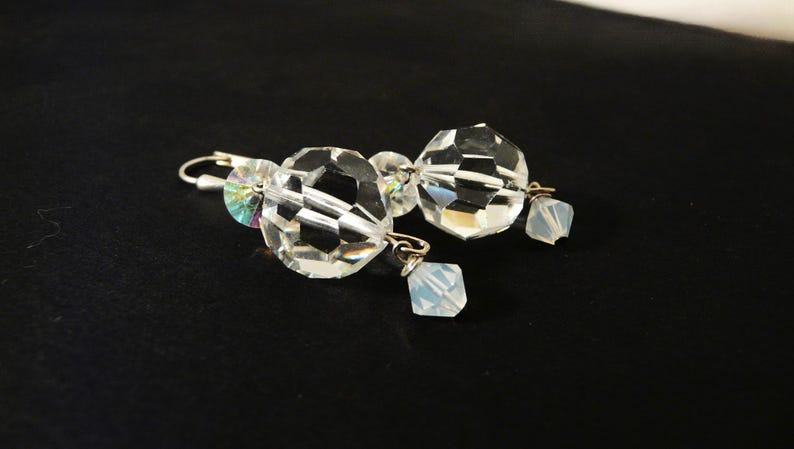 May include: A pair of clear crystal earrings with faceted, spherical and square-shaped beads. The earrings have silver-colored hooks and are displayed against a black background. The crystals reflect light, creating a sparkling effect.