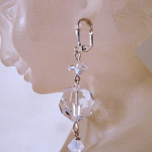 May include: A single, dangling earring with a silver-colored clasp. The earring features a clear, faceted crystal sphere and a smaller, square crystal, connected by delicate silver chains. The crystals reflect light, creating a sparkling effect.
