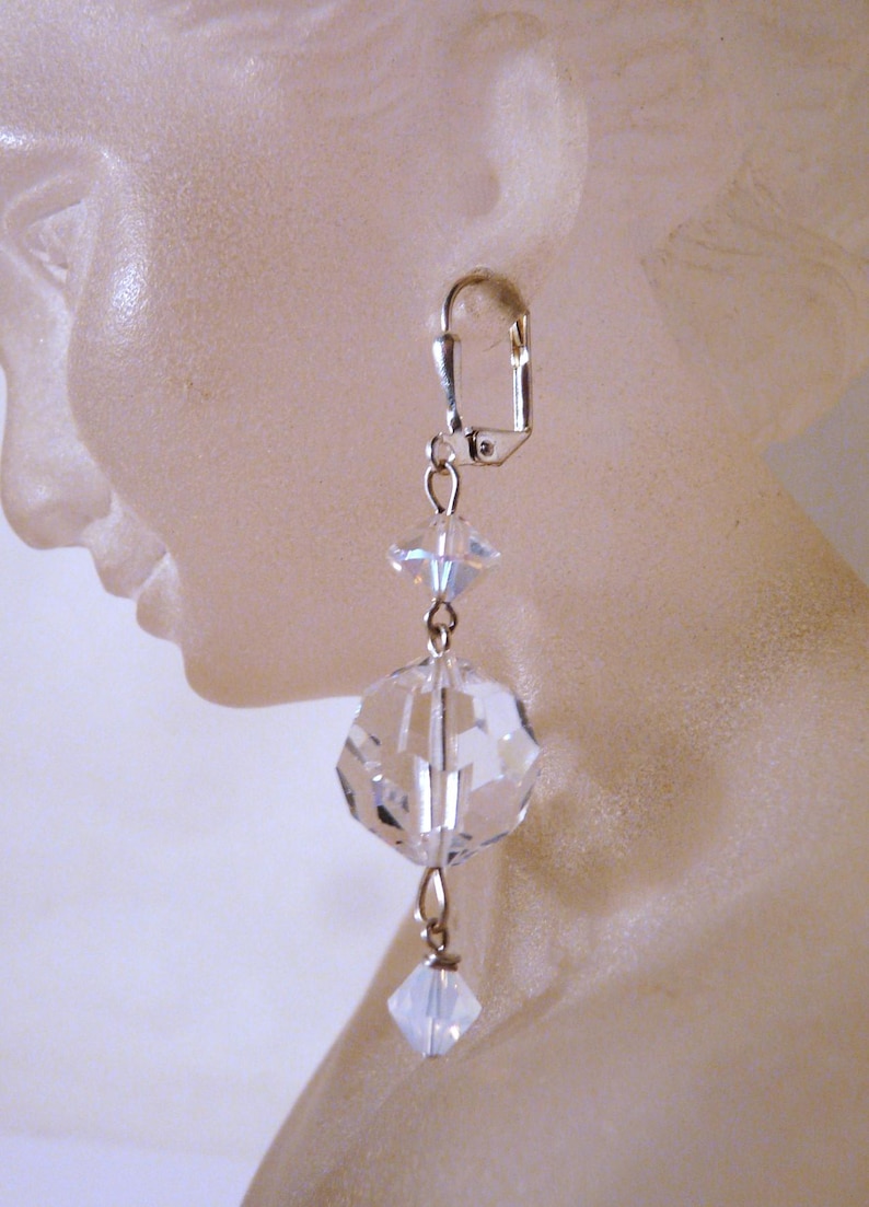 May include: A single, dangling earring with a silver-colored clasp. The earring features a clear, faceted crystal sphere and a smaller, square crystal, connected by delicate silver chains. The crystals reflect light, creating a sparkling effect.