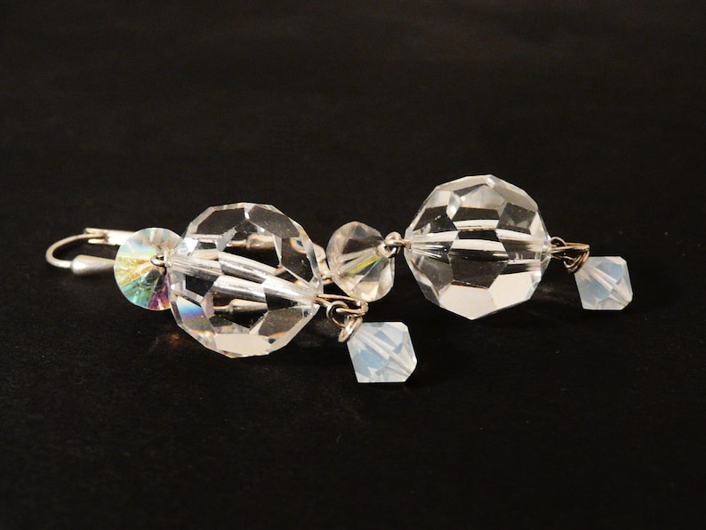 May include: Clear crystal earrings with faceted, spherical and square-shaped beads. The earrings have a silver-colored hook closure and small iridescent accents. The jewelry is displayed against a dark background.