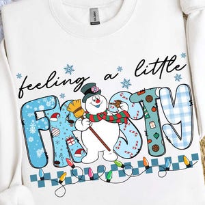 May include: White sweatshirt with the phrase "feeling a little frosty" and a snowman graphic. The word "frosty" is in blue, with snowflakes and holiday-themed patterns. A string of colorful lights decorates the bottom.