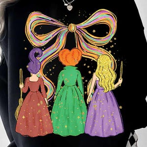May include: Black sweatshirt featuring a colorful bow design and a graphic of three cartoon witches in dresses. The witches are in red, green, and purple dresses, with gold glitter accents. One witch holds a broom, and another holds a wand.