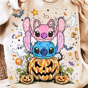 May include: Beige sweatshirt featuring a Halloween-themed graphic. The design includes a blue and pink cartoon character duo atop a carved pumpkin, surrounded by ghosts, bats, stars, and candy. The sweatshirt has a crew neck and long sleeves.