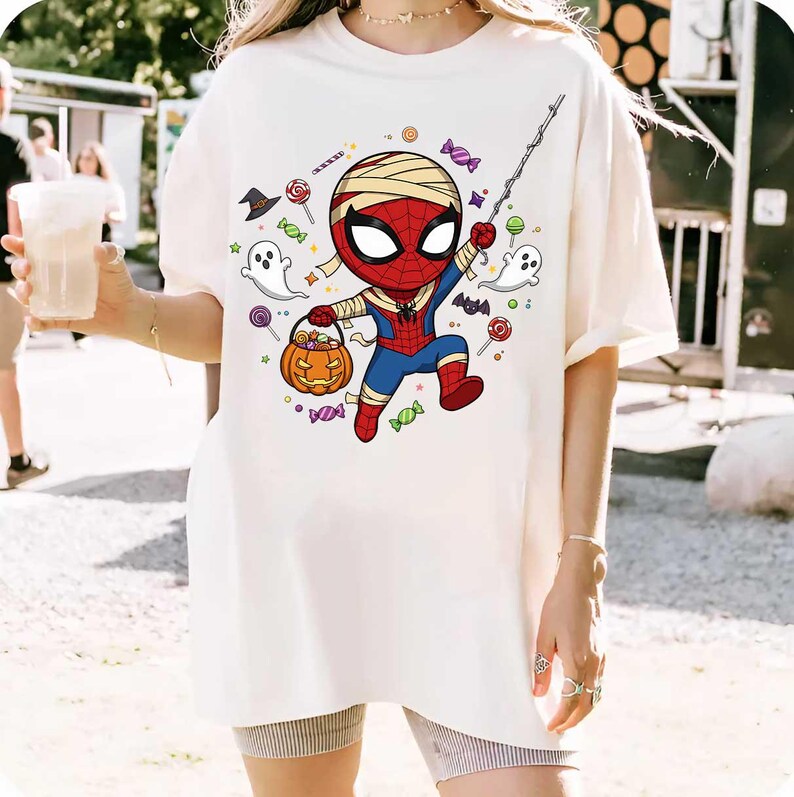 May include: White t-shirt featuring a cartoon Spider-Man character dressed as a mummy holding a pumpkin basket. The design includes Halloween candy, ghosts, and a spider web. The shirt is worn by a person.