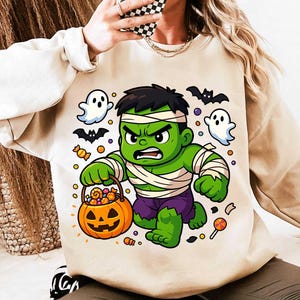 May include: Beige sweatshirt featuring a cartoon Hulk character dressed as a mummy holding a jack-o'-lantern candy bucket. The design includes ghosts, bats, and candy, perfect for Halloween.