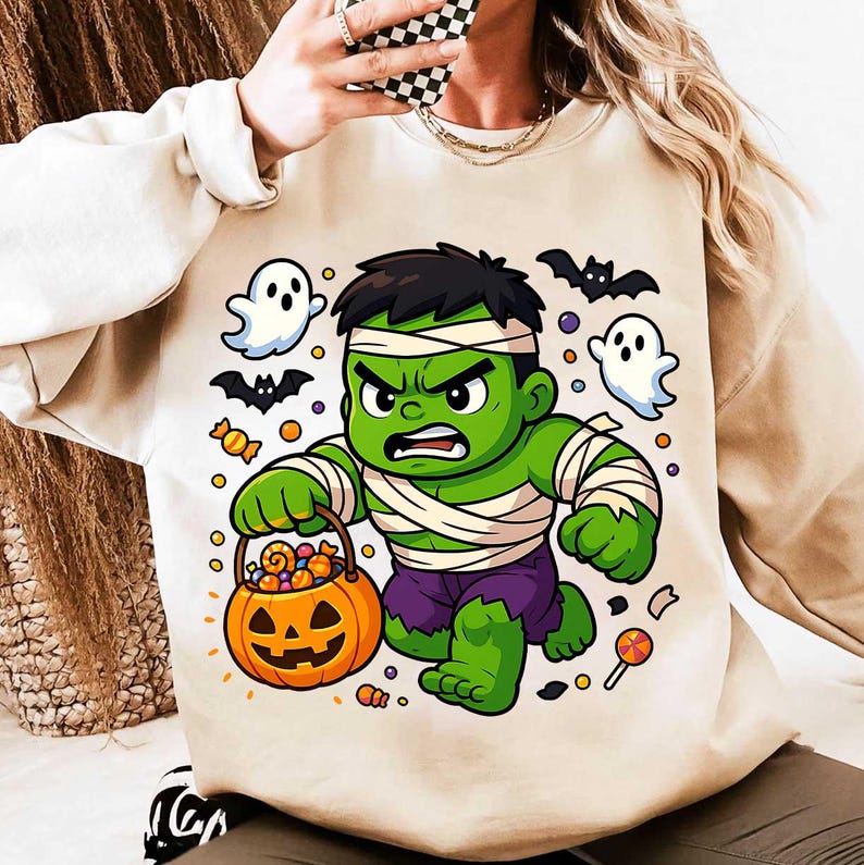 May include: Beige sweatshirt featuring a cartoon Hulk character dressed as a mummy holding a jack-o'-lantern candy bucket. The design includes ghosts, bats, and candy, perfect for Halloween.